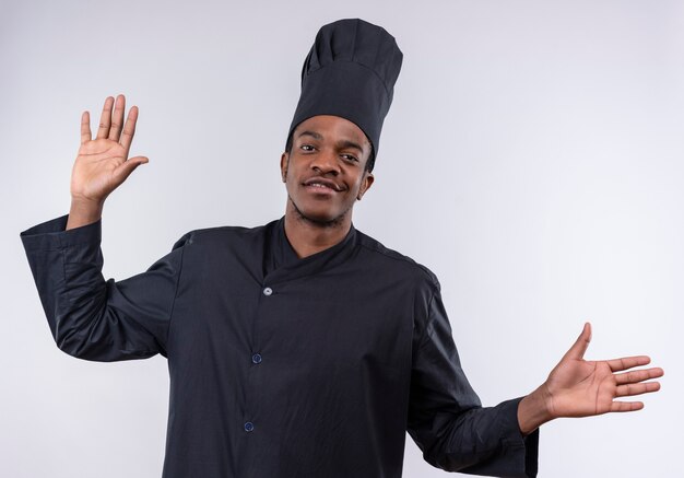 Young confident afro-american cook in chef uniform raises hands up isolated on white background with copy space