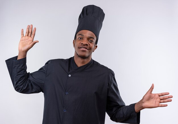 Молодой уверенный в себе афро-американский повар в униформе шеф-повара поднимает руки вверх на белом фоне с копией пространства