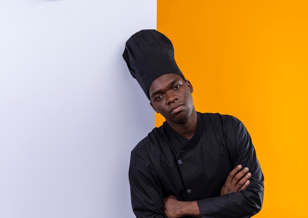 Free photo young confident afro-american cook in chef uniform stands in front of white wall with crossed arms on orange  with copy space