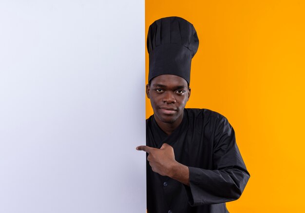 Young confident afro-american cook in chef uniform stands behind and points at white wall isolated on orange space with copy space