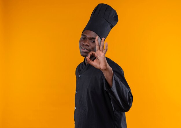 요리사 제복을 입은 젊은 자신감 아프리카 계 미국인 요리사가 옆으로 서서 복사 공간이있는 오렌지에 확인 손 기호를 제스처