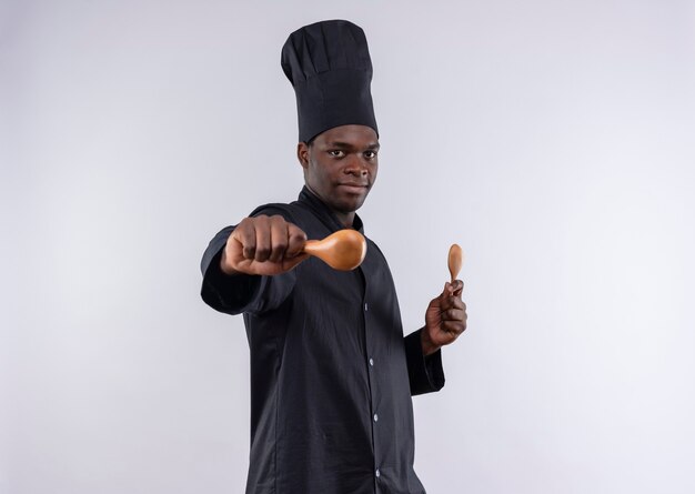 Young confident afro-american cook in chef uniform stands sideways and holds wooden spoons on white  with copy space