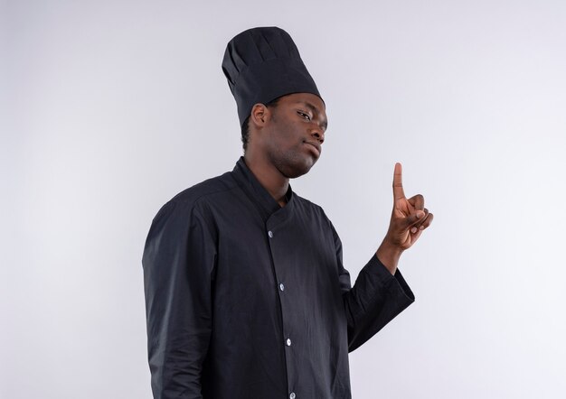 Молодой уверенный в себе афроамериканский повар в униформе шеф-повара стоит боком и указывает на белый с копией пространства