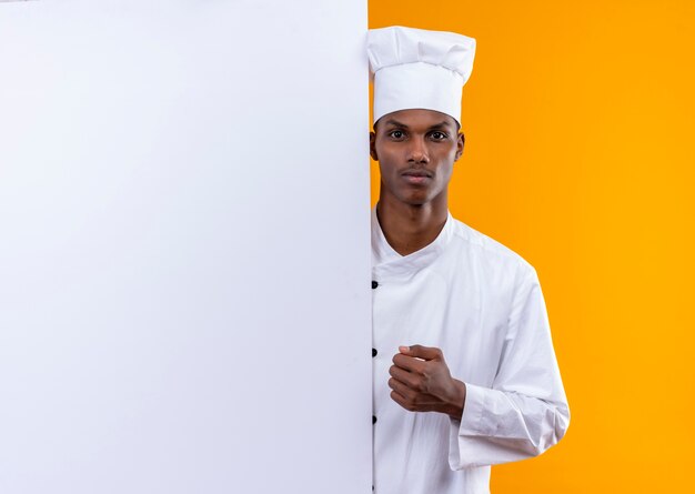 Молодой уверенный в себе афро-американский повар в униформе шеф-повара стоит за белой стеной и держит кулак на оранжевом фоне с копией пространства