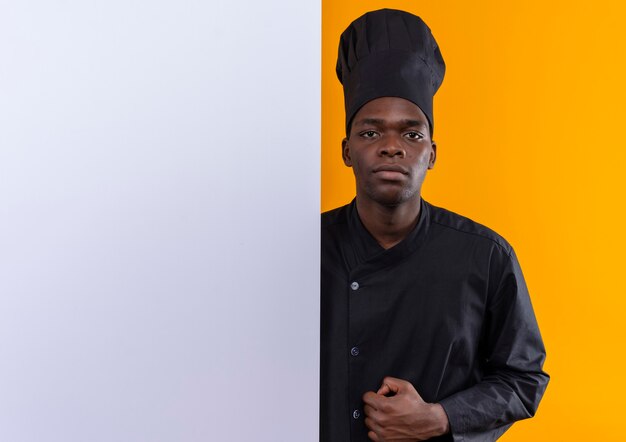 Young confident afro-american cook in chef uniform stands behind white wall on orange  with copy space