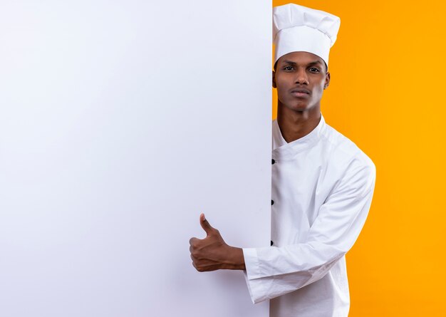 Молодой уверенный в себе афро-американский повар в униформе шеф-повара стоит за белой стеной и показывает палец вверх на оранжевом фоне с копией пространства