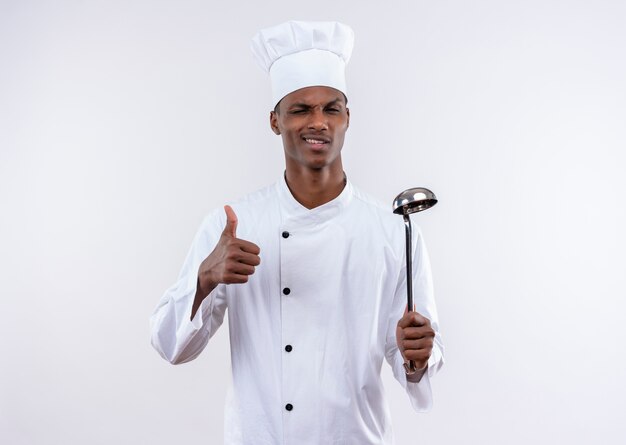 Young confident afro-american cook in chef uniform thumbs up blinks with eye and holds ladle isolated on white background with copy space