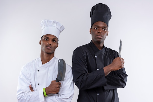 Young confident afro-american cooks in chef uniform with crossed arms hold knifes isolated on white background