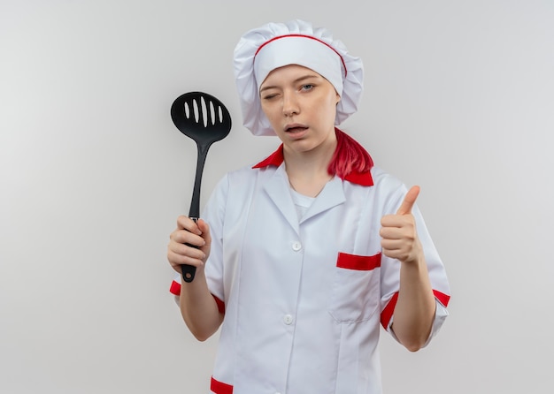 Il giovane chef femminile biondo sicuro in uniforme del cuoco unico lampeggia gli occhi e il pollice in alto tenendo la spatola isolata sul muro bianco