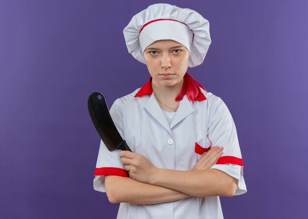 Young confident blonde female chef in chef uniform crosses arms and holds knife isolated on violet wall