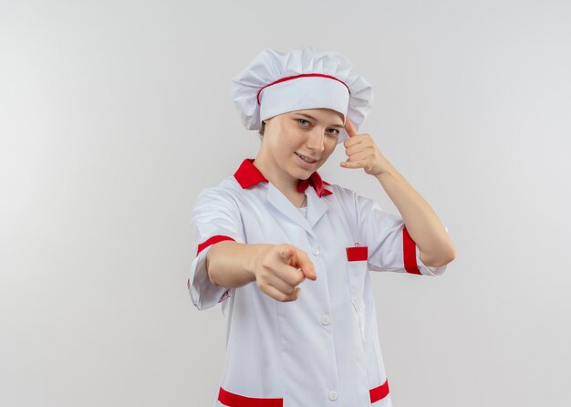 Young confident blonde female chef in chef uniform gestures call me hand sign and points isolated on white wall