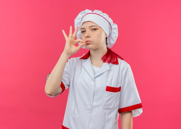 Young confident blonde female chef in chef uniform gestures delicious sign isolated on pink wall