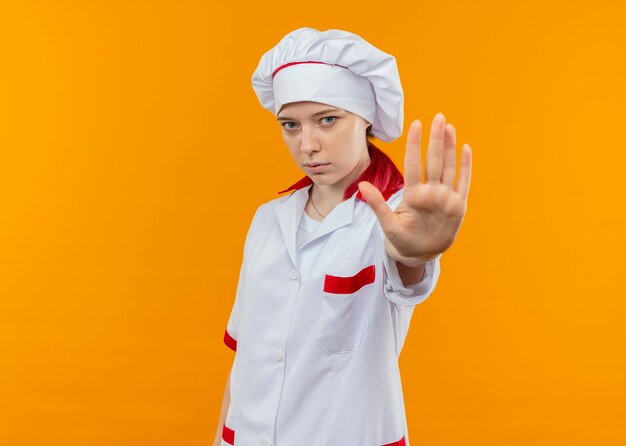 Young confident blonde female chef in chef uniform gestures stop hand sign isolated on orange wall