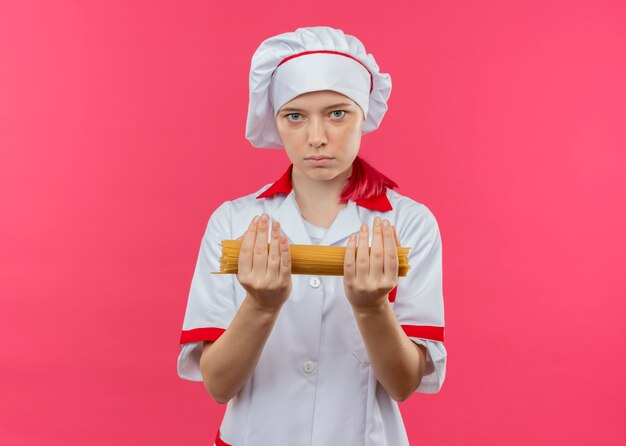 Молодая уверенная в себе блондинка шеф-повар в униформе шеф-повара держит кучу спагетти обеими руками, изолированными на розовой стене