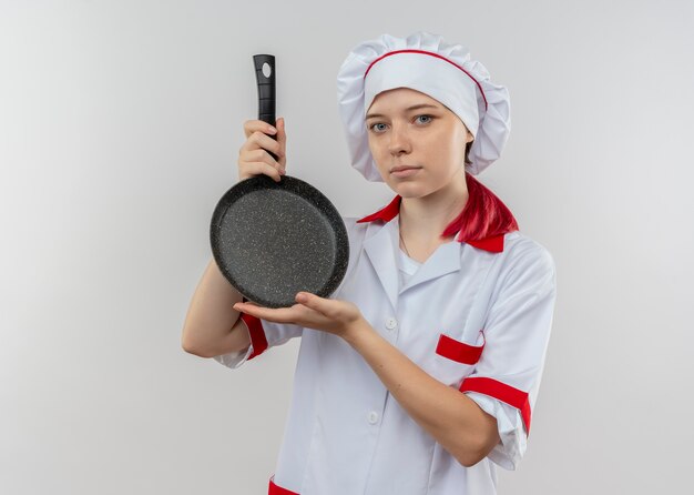 Young confident blonde female chef in chef uniform holds frying pan with two hands isolated on white wall