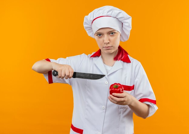 Young confident blonde female chef in chef uniform holds knife and red pepper isolated on orange wall