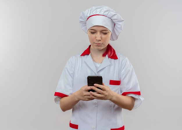 Молодая уверенная блондинка женщина-повар в форме шеф-повара держит и смотрит на телефон, изолированный на белой стене