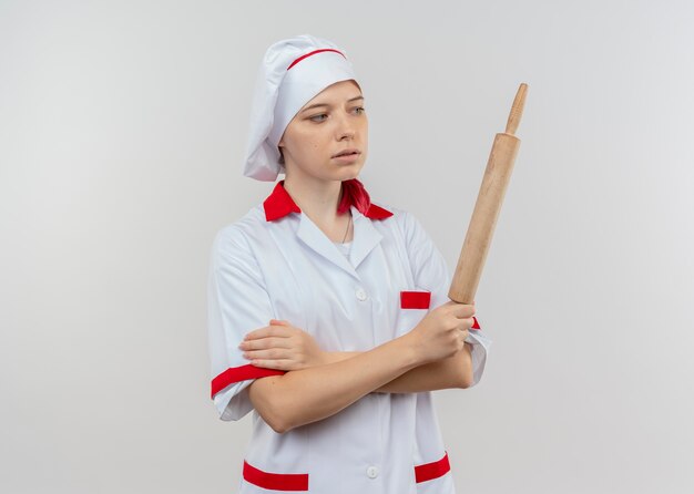 Young confident blonde female chef in chef uniform holds and looks at rolling pin isolated on white wall