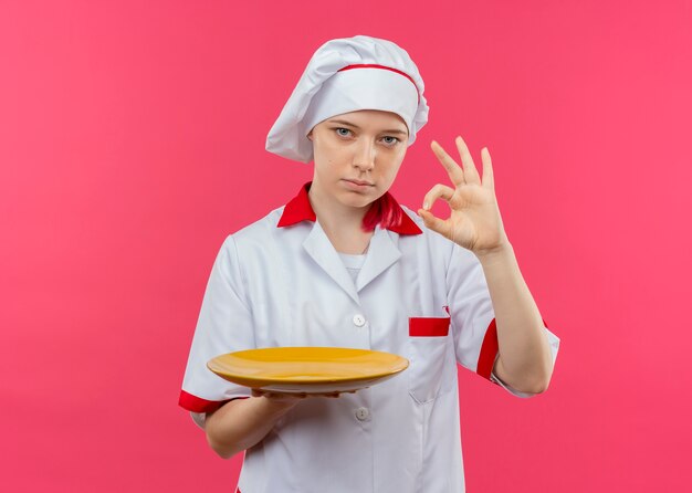 Молодая уверенная блондинка-шеф-повар в униформе шеф-повара держит тарелку и жестикулирует знак рукой на розовой стене