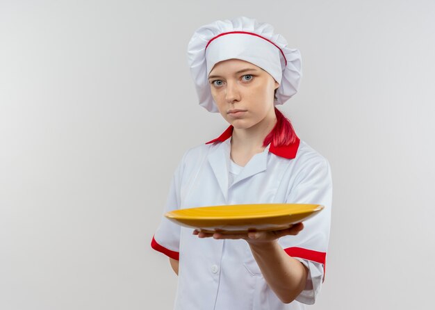 Young confident blonde female chef in chef uniform holds plate and keeps hand behind isolated on white wall