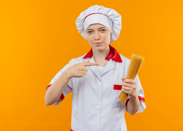 Young confident blonde female chef in chef uniform holds and points at bunch of spaghetti isolated on orange wall