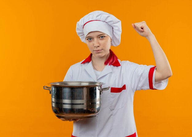 Young confident blonde female chef in chef uniform holds pot and raises fist up isolated on orange wall