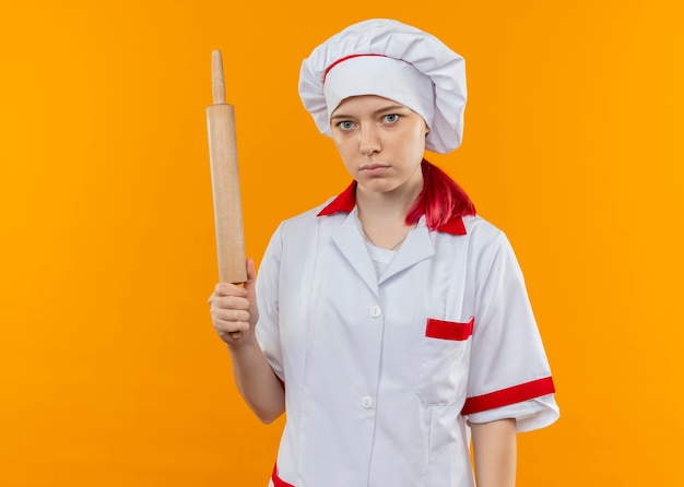 Young confident blonde female chef in chef uniform holds rolling pin isolated on orange wall