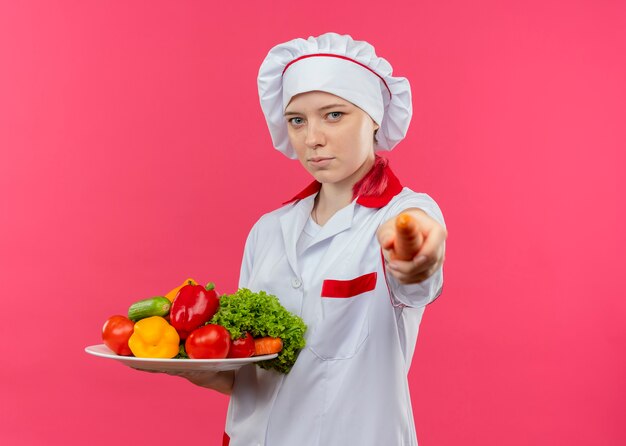 요리사 제복을 입은 젊은 자신감 금발 여성 요리사는 접시에 야채를 보유하고 분홍색 벽에 고립 된 당근을 보유하고 있습니다.