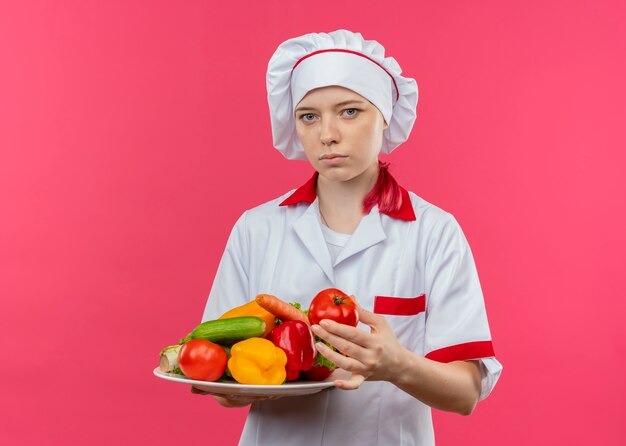 Young confident blonde female chef in chef uniform holds vegetables on plate and looks isolated on pink wall