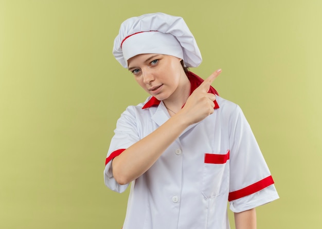 Young confident blonde female chef in chef uniform points at side and looks isolated on green wall