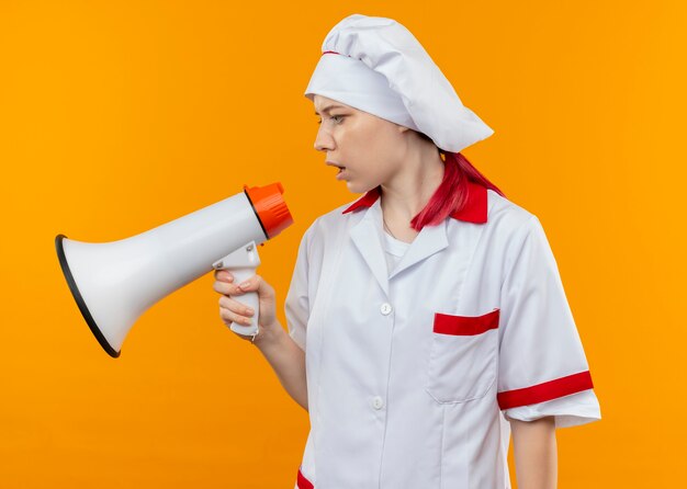 Young confident blonde female chef in chef uniform speaks through loud speaker and looks at side isolated on orange wall