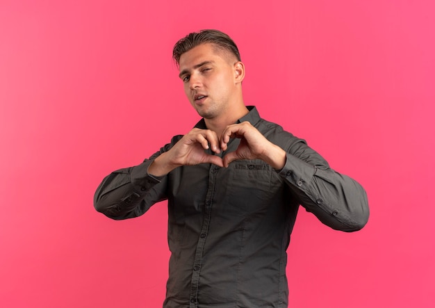 Free photo young confident blonde handsome man gestures heart hand sign looking at camera isolated on pink background with copy space