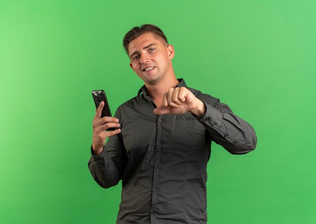 Young confident blonde handsome man holds and points at phone isolated on green background with copy space