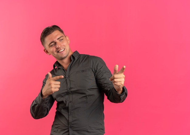 Young confident blonde handsome man points and looks at side isolated on pink background with copy space