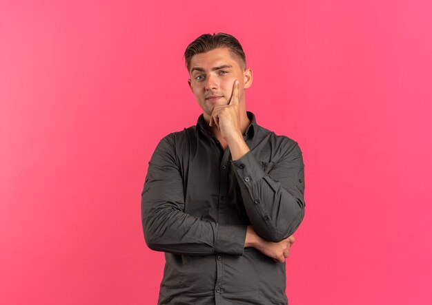 Young confident blonde handsome man puts hand on chin looking at camera isolated on pink background with copy space