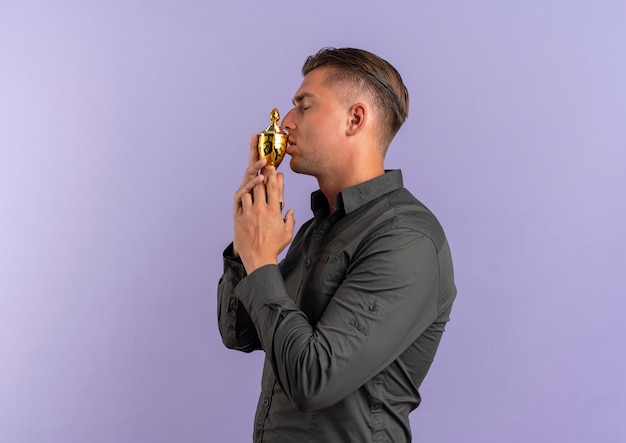 Young confident blonde handsome man stands sideways and holds cup isolated on violet space with copy space