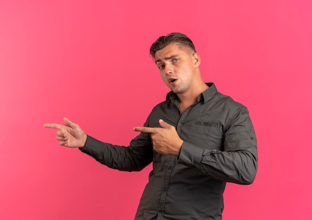 Free photo young confident blonde handsome manpoints at side looking at camera isolated on pink background with copy space