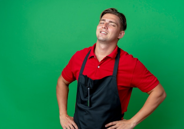 young confident blonde male barber in uniform blinks eye puts hands on waist