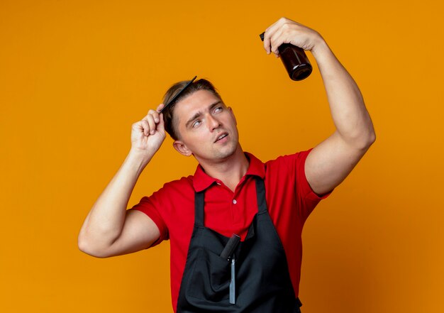 Free photo young confident blonde male barber in uniform combing hair looking at spray bottle isolated on orange space with copy space