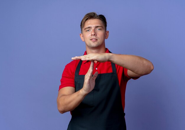 Молодой уверенный в себе светловолосый мужчина-парикмахер в униформе жесты, тайм-аут, знак рукой, изолированный на фиолетовом пространстве с копией пространства
