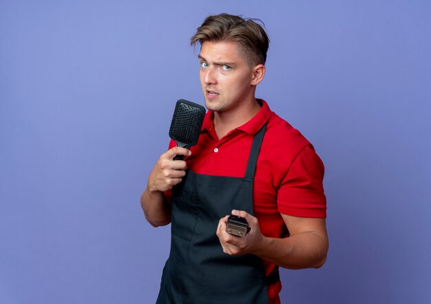 Young confident blonde male barber in uniform holds comb and hair clipper isolated on violet space with copy space