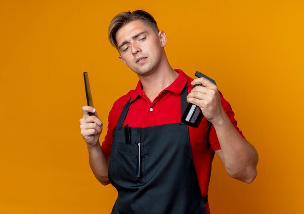 Young confident blonde male barber in uniform holds comb and looks at spray bottle isolated on orange space with copy space