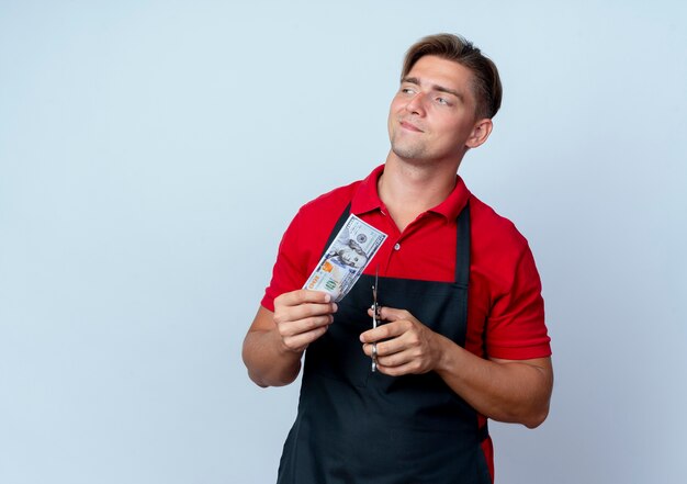 Young confident blonde male barber in uniform holds hundred dollars bill and scissors looking at side isolated on white space with copy space