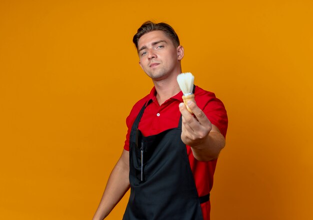 Young confident blonde male barber in uniform holds out shaving brush isolated on orange space with copy space