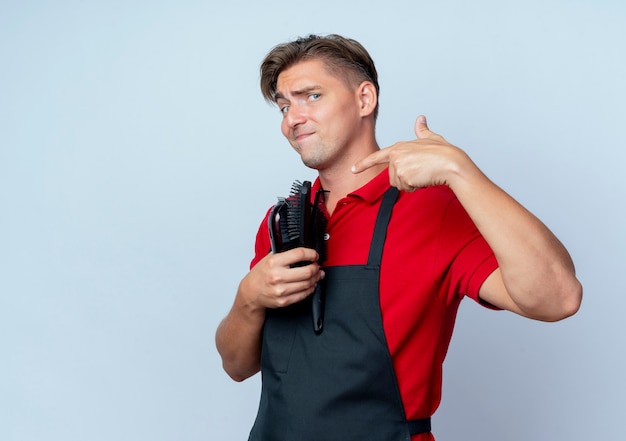 Young confident blonde male barber in uniform holds and points at barber tools isolated on white space with copy space