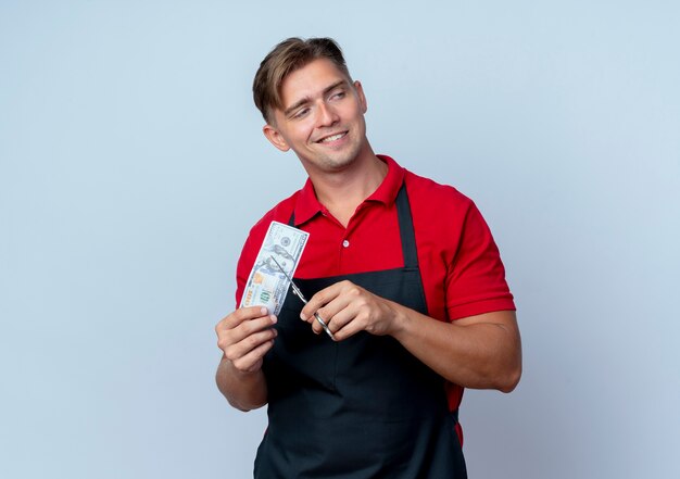 Young confident blonde male barber in uniform pretends to cut hundred dollars bill looking at side isolated on white space with copy space