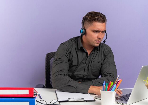 Young confident blonde office worker man on headphones sits at desk with office tools looking at laptop isolated on violet background with copy space