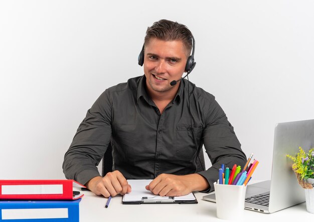헤드폰에 젊은 자신감 금발 회사원 남자는 복사 공간 흰색 배경에 고립 된 카메라에서 노트북을 사용하는 사무실 도구와 책상에 앉아