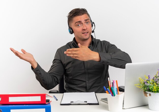 헤드폰에 젊은 자신감 금발 회사원 남자 복사 공간이 흰색 배경에 고립 된 손으로 측면에서 노트북 포인트를 사용하여 사무실 도구와 책상에 앉아