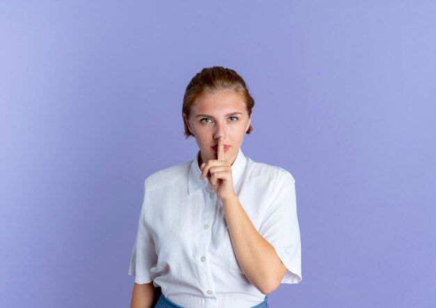 Young confident blonde russian girl gestures hush quiet isolated on purple background with copy space